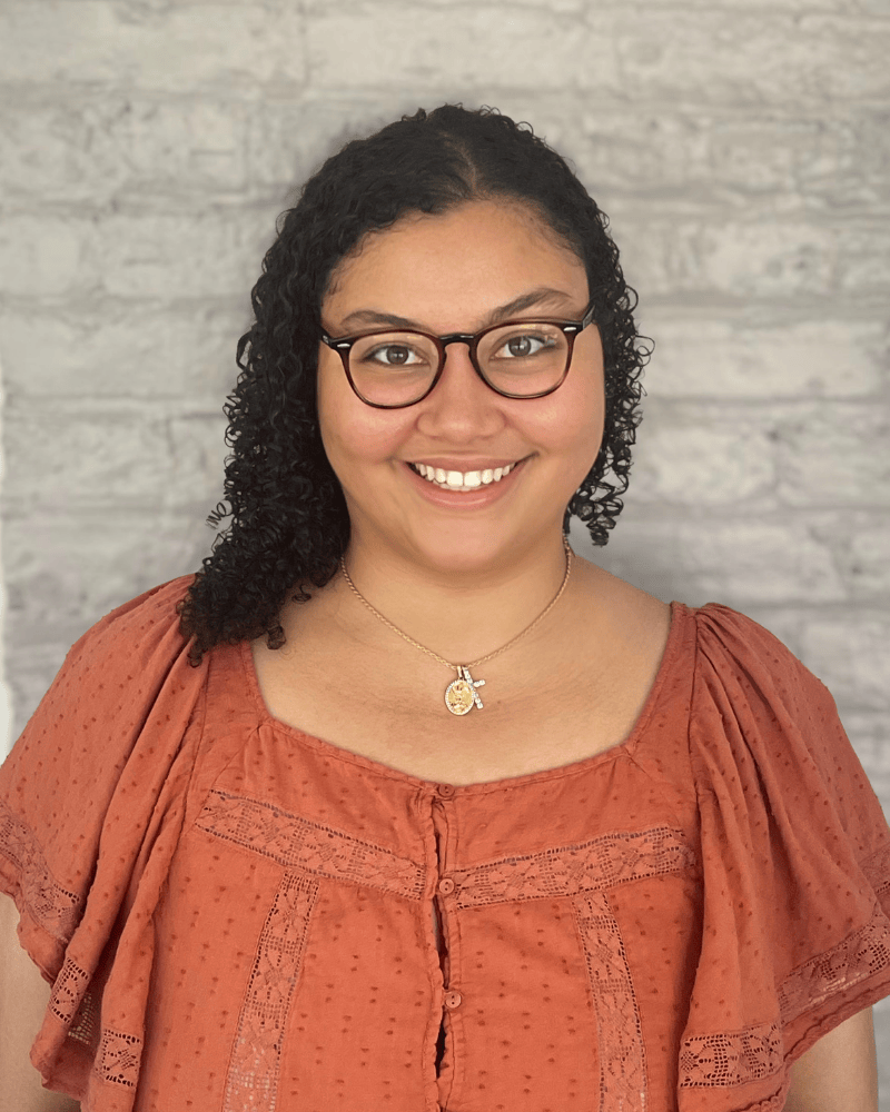 Portrait of a smiling woman with curly dark hair and glasses wearing a rust-colored blouse and necklace, standing in front of a light brick wall.